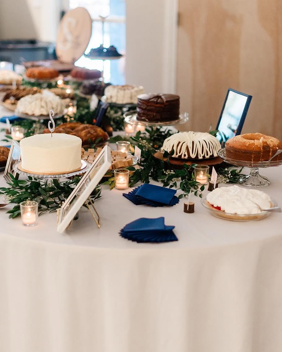 A dessert table draped in white linen against a wood-paneled wall. Greenery and lit votives run between cakes and pies on glass stands and wooden boards, including a white frosted cake with a gold letter topper, a chocolate layer cake, a bundt cake with white icing, and a pie topped with white meringue. Navy napkins are stacked on the cloth, and a framed sign leans at the front.