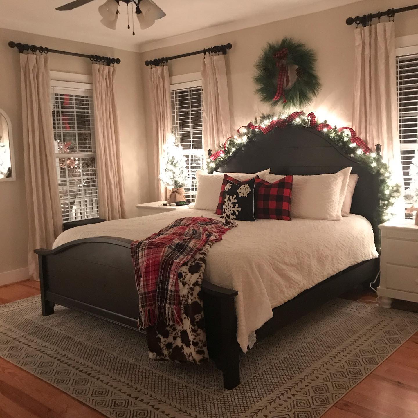 A bedroom with a dark wood bed, a white quilt, and a red plaid throw. A black pillow with a white snowflake and a red-and-black plaid pillow sit against white pillows. A green garland with white lights and red plaid bows runs along the headboard, and a green wreath with a red plaid bow hangs above it. Cream curtains frame the windows. A small lit tree stands on a nightstand. A patterned rug covers the wood floor.